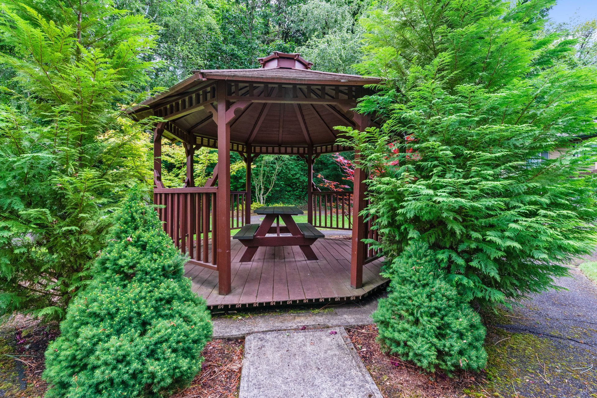 Wooden gazebo with picnic table surrounded by lush green trees and shrubs in a garden path setting