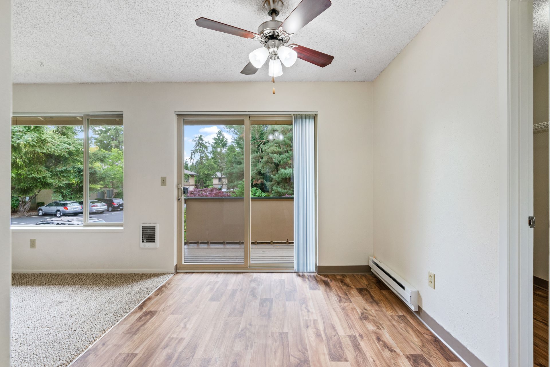 Empty living room with wood floor, ceiling fan, sliding glass door, and large windows overlooking greenery