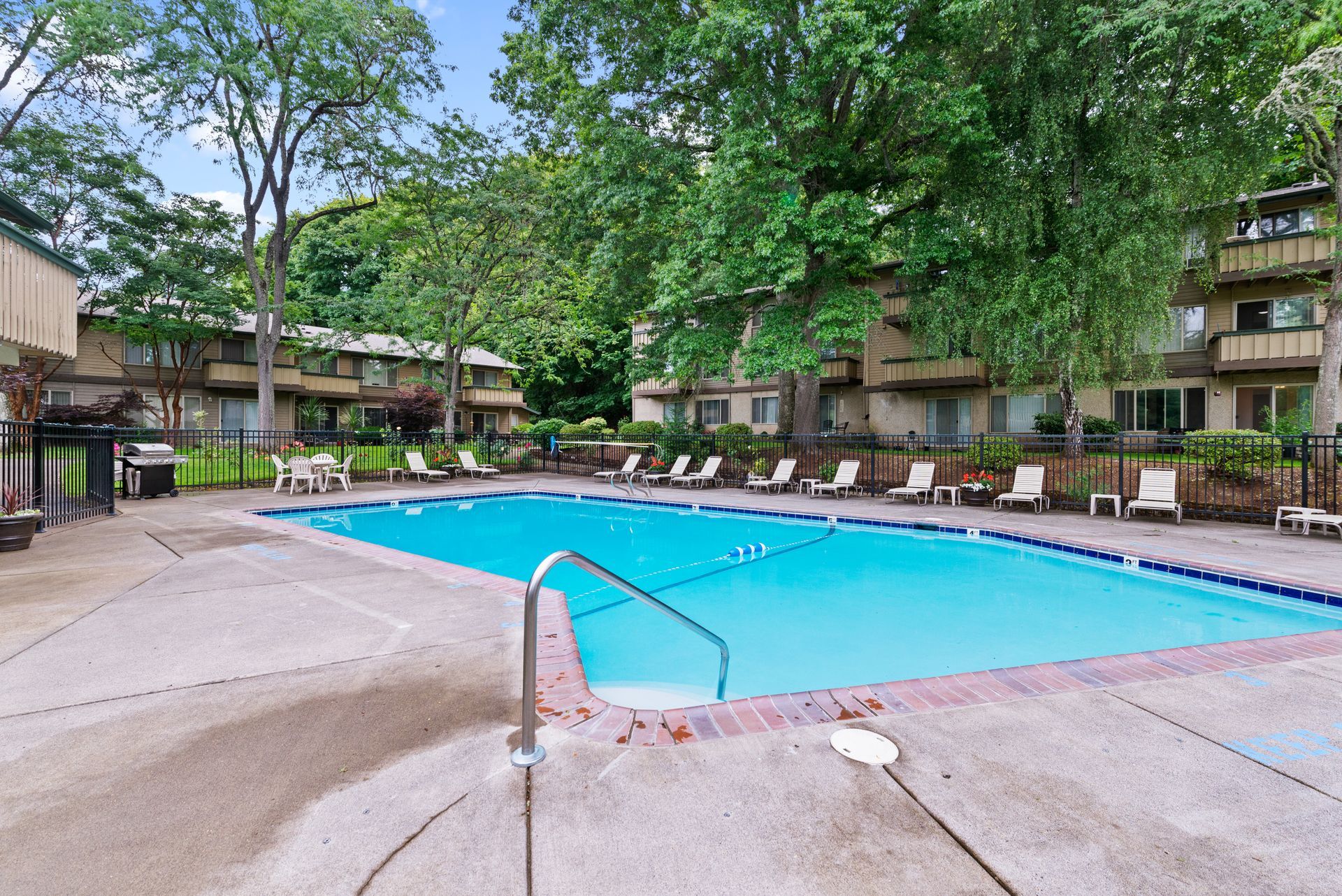 Outdoor apartment pool with turquoise water, surrounded by trees and concrete deck.