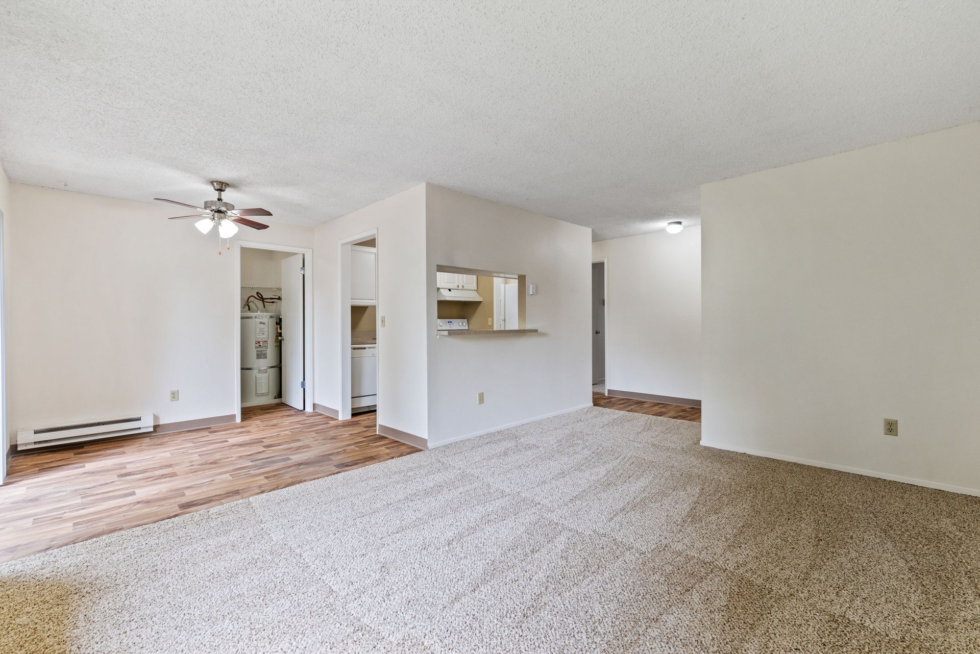 Empty beige living room with carpet, wood flooring, ceiling fan, and pass-through kitchen opening
