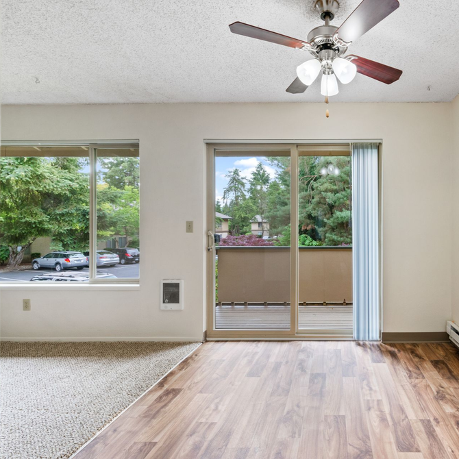 Empty living room with wood floor, ceiling fan, large window, and sliding glass door to a balcony.