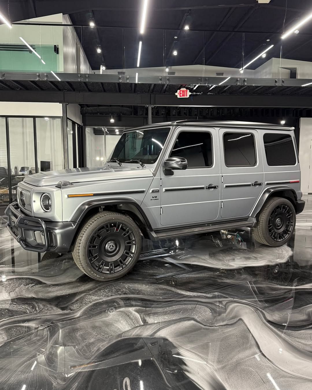 Silver Mercedes G-Class SUV parked on a shiny black and white floor under bright lights.