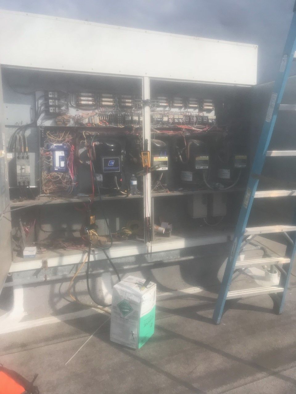 Electrical service panel with open wiring, a blue ladder, and a paint bucket on a concrete floor outdoors