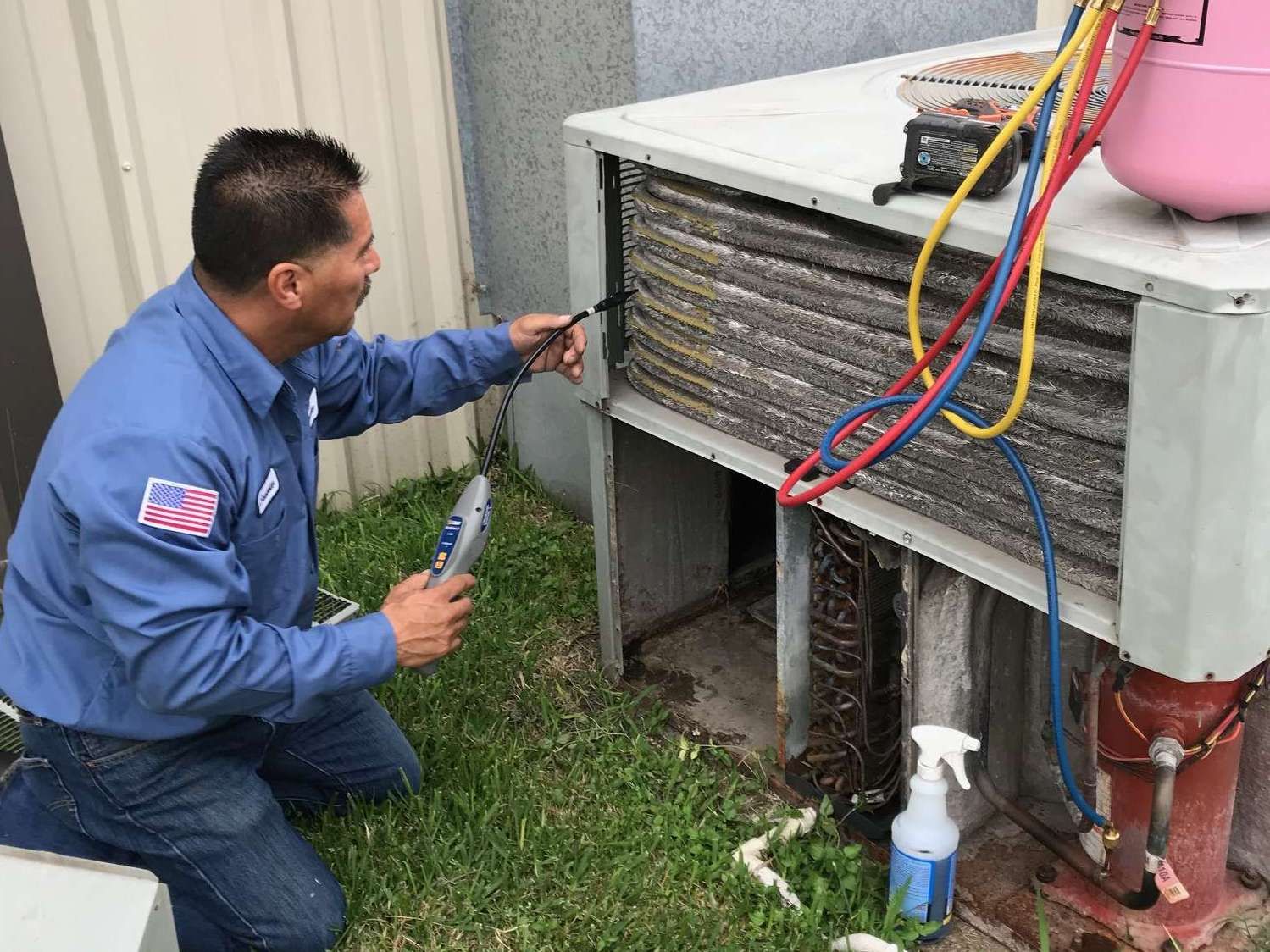 Technician kneeling beside an outdoor HVAC unit, inspecting coils with colored hoses attached.