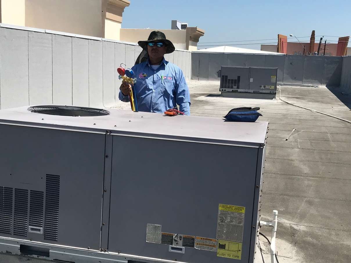 Worker in blue shirt and hard hat standing on a rooftop beside HVAC units, holding a tool.
