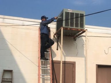 Worker on a ladder servicing an outdoor air conditioner unit mounted on a building wall.