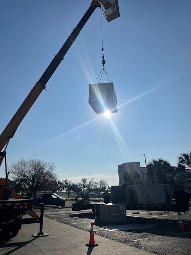 Crane lifting a large metal bin on a sunny construction site, with sun glare in the sky.