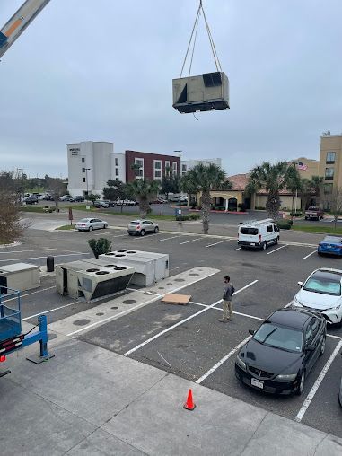 Crane lifting a small structure over a parking lot, with a worker and traffic cones below.