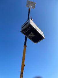 A yellow telescoping lift raises a large solar-powered device against a blue sky.