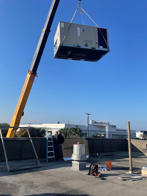 Crane lifting a large HVAC unit over a rooftop construction site under a clear blue sky