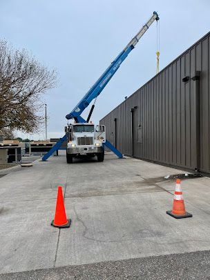 Crane truck with extended blue boom beside a warehouse, blocked by orange cones.