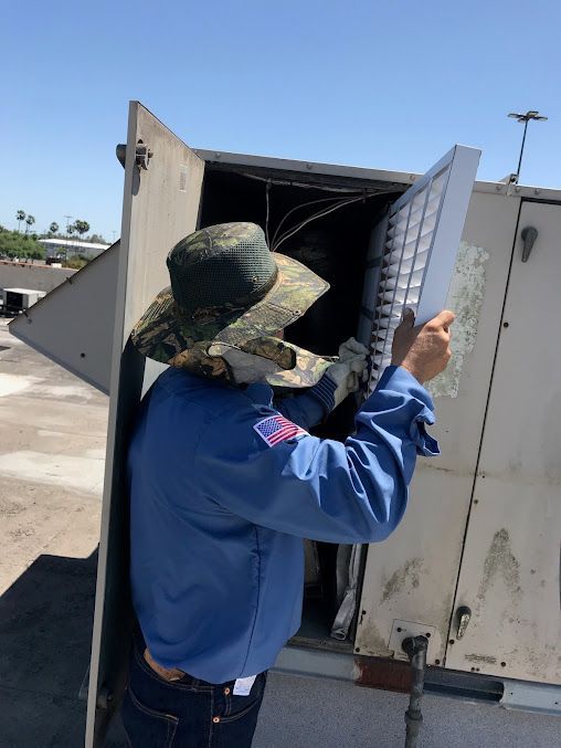 Worker in blue shirt and sun hat inspecting an open utility unit outdoors.