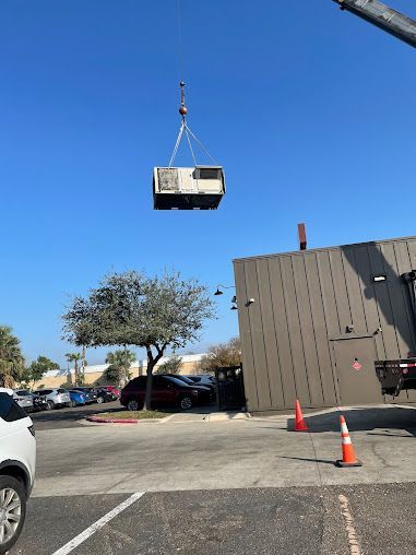 Crane lifting a large HVAC unit above a parking lot beside a gray building and orange cones