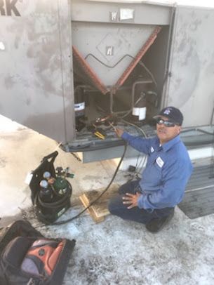 Technician kneeling by an open HVAC unit in snow, inspecting wiring with tools and gas tanks nearby