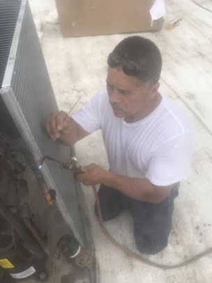 Man in a white T-shirt kneels beside outdoor HVAC unit, working with a tool on the metal panel.