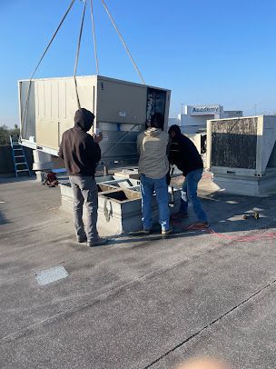 Three workers guide a large HVAC unit being lifted by crane on a rooftop.