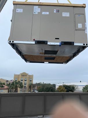 Large beige HVAC unit suspended by a crane above a building roof under a cloudy sky