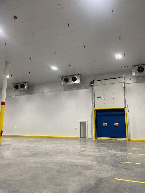 Large industrial warehouse interior with white walls, concrete floor, blue loading dock door, and overhead fans.