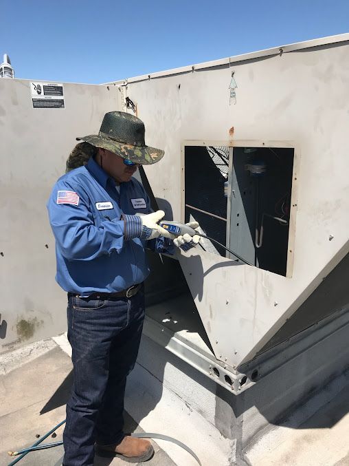 Worker in blue uniform inspects equipment on a rooftop, using tools near an open metal panel.