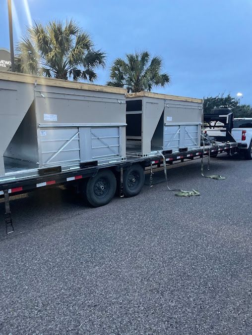 Flatbed truck carrying two gray shipping containers on a street with palm trees nearby