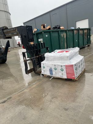 Forklift beside a pallet of wrapped goods near green dumpsters on a wet loading dock