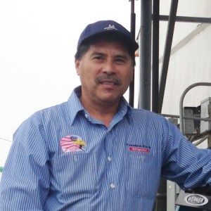 Man in blue striped shirt and cap, standing outdoors near industrial equipment.