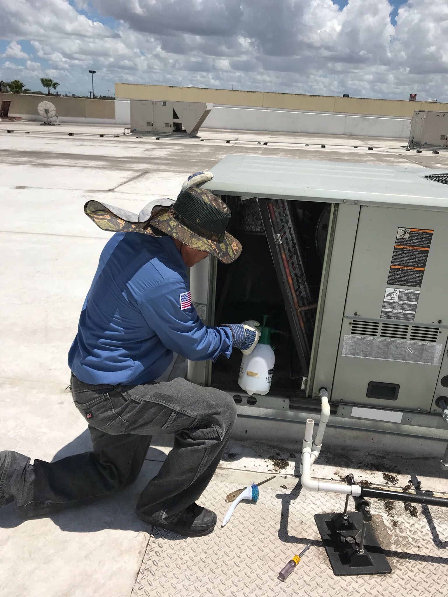 Technician kneels beside rooftop HVAC unit, inspecting piping under a bright blue sky.