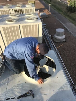 Worker kneeling on a rooftop repairing a round HVAC vent beside large AC units.