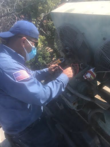 Technician in blue uniform and mask inspecting wiring inside an open vehicle hood outdoors.