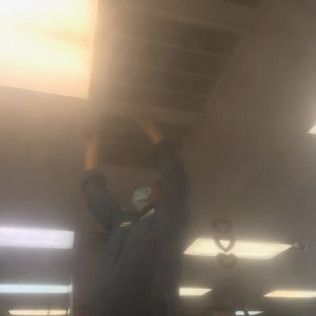 Person reaching up toward a ceiling fixture in a dim indoor room, wearing a mask.