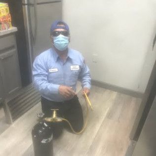 Person in a blue uniform and mask kneeling beside a fire extinguisher in a room, holding a cable or tool.