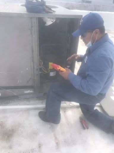 Technician in blue uniform inspecting electrical equipment with a tool on a snowy rooftop.
