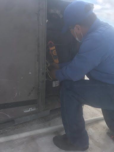Technician kneels beside an outdoor HVAC unit, inspecting wiring with a tool.