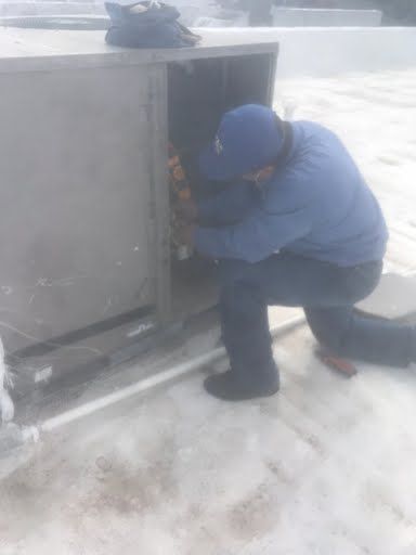 Person in blue jacket crouches beside a rooftop HVAC unit in snowy conditions.
