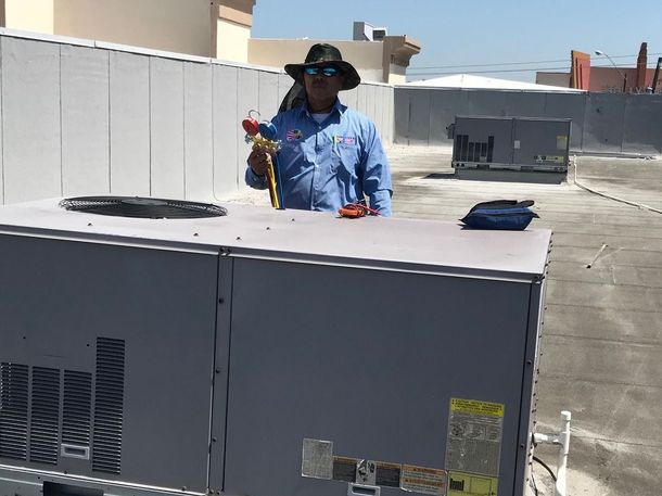 Worker in blue shirt and hat standing on a rooftop beside HVAC units, holding a tool.
