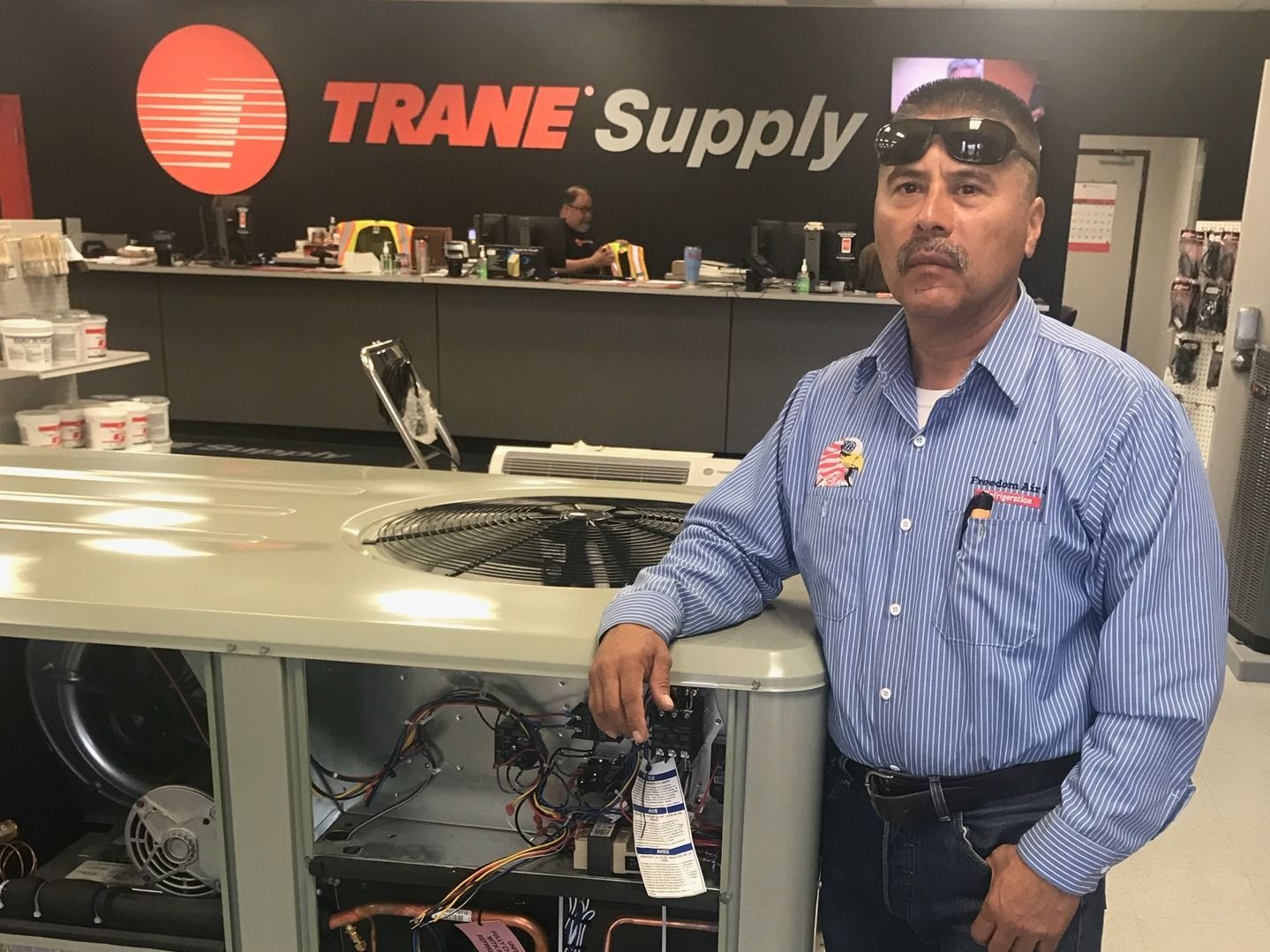 Man standing beside a Trane HVAC unit at a trade show booth, with the TRANE Supply sign in the background.