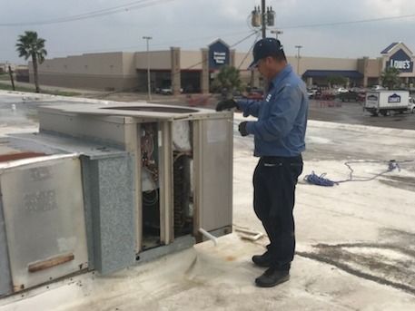 Worker in blue jacket inspecting rooftop HVAC unit beside a Lowe’s store.