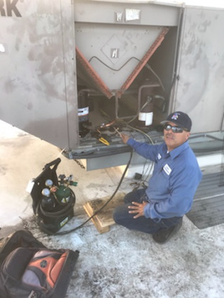 Technician kneels beside open HVAC unit, inspecting wiring on a snowy rooftop.