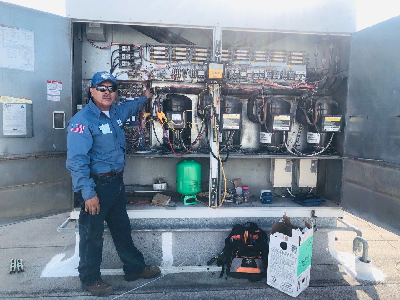 Technician in blue uniform standing beside an open electrical panel with tools and equipment outdoors