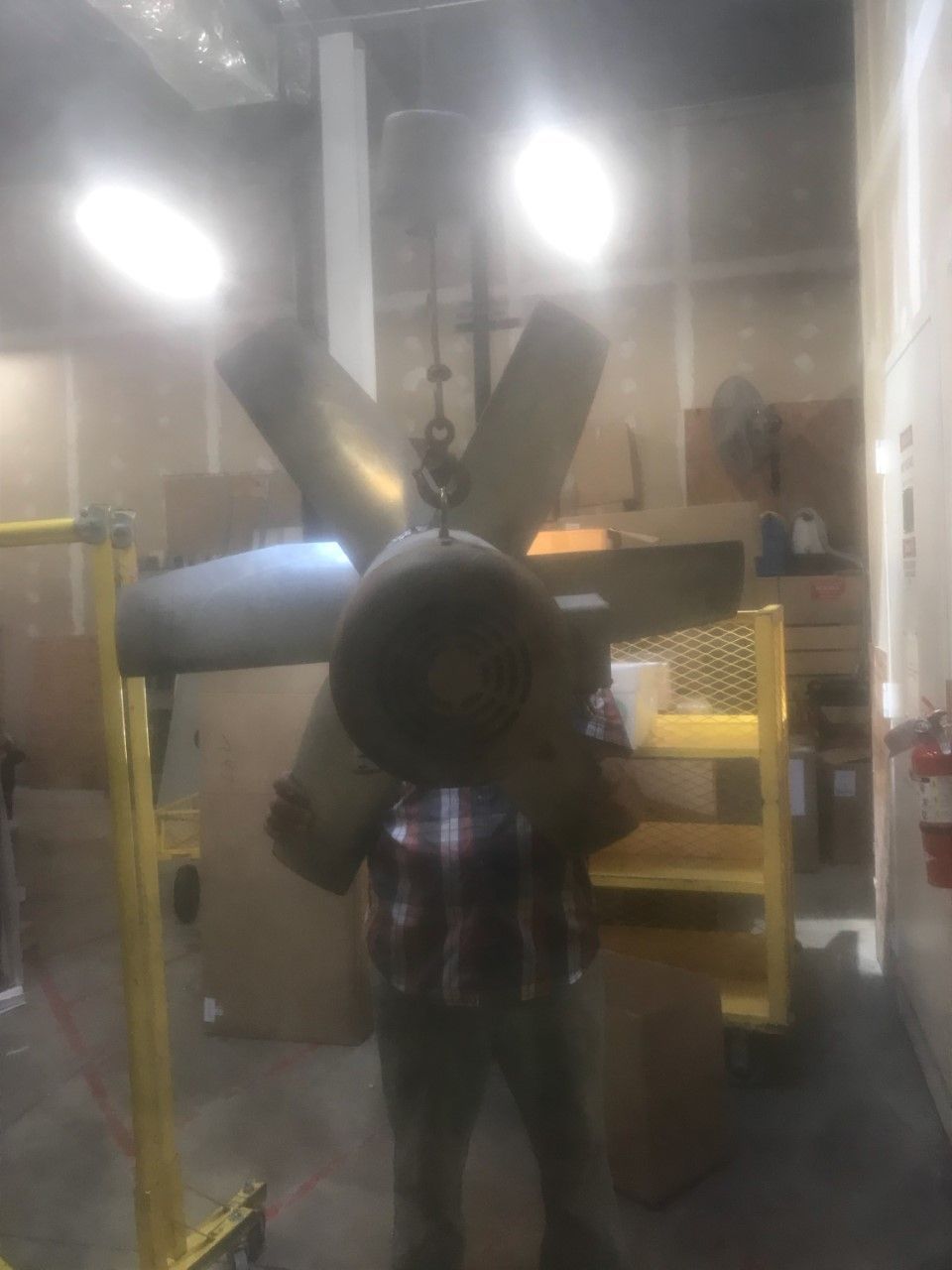 Person standing in a workshop beside large metal ducts and machinery under bright lights.