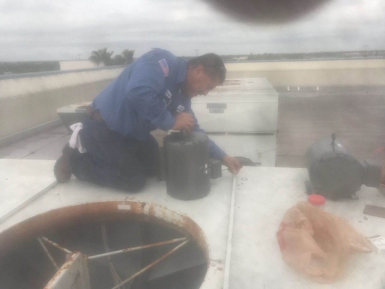 Worker crouching on a rooftop beside black equipment and a tan bag near a large vent opening.