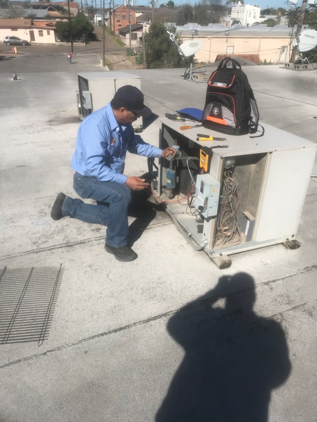 Technician kneels beside rooftop HVAC unit, inspecting exposed components with tools.