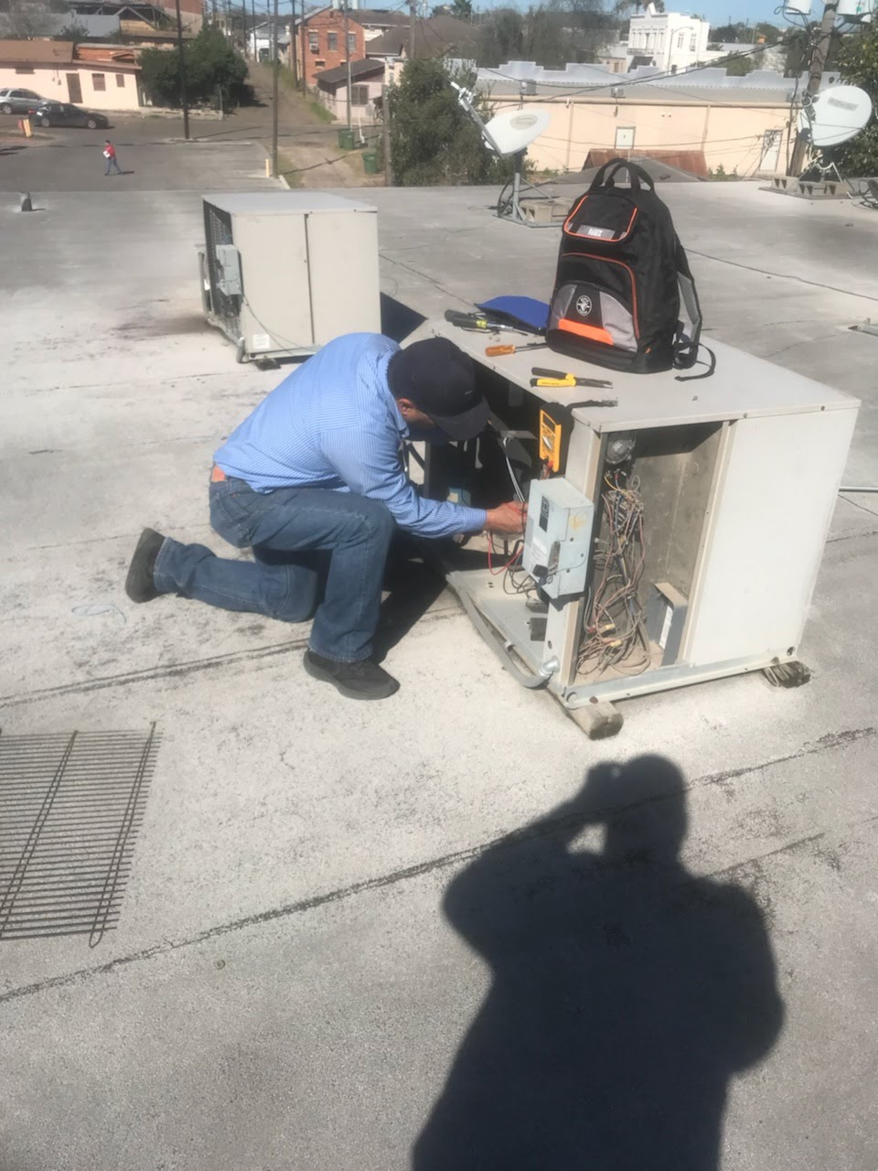 Technician crouches beside an outdoor HVAC unit, repairing wiring on a concrete rooftop.