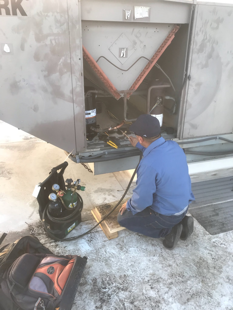 Worker kneels beside open HVAC unit, repairing wiring on a snowy rooftop.