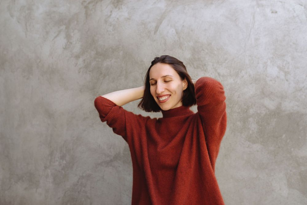 Una donna con un maglione rosso è in piedi davanti a un muro grigio con le mani dietro la testa.