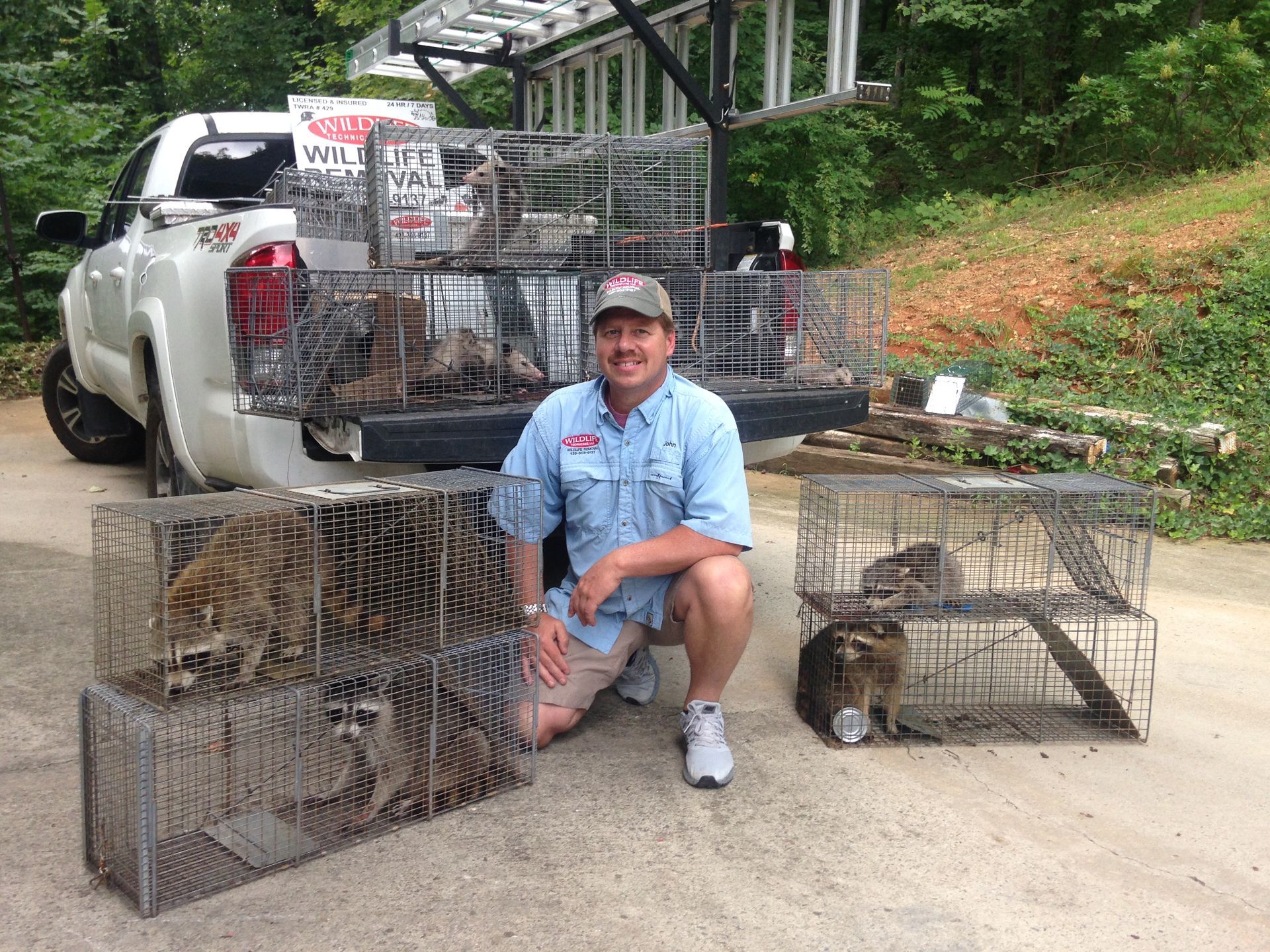 Man kneels by live animal traps beside a white truck on a driveway, with more traps stacked behind him.