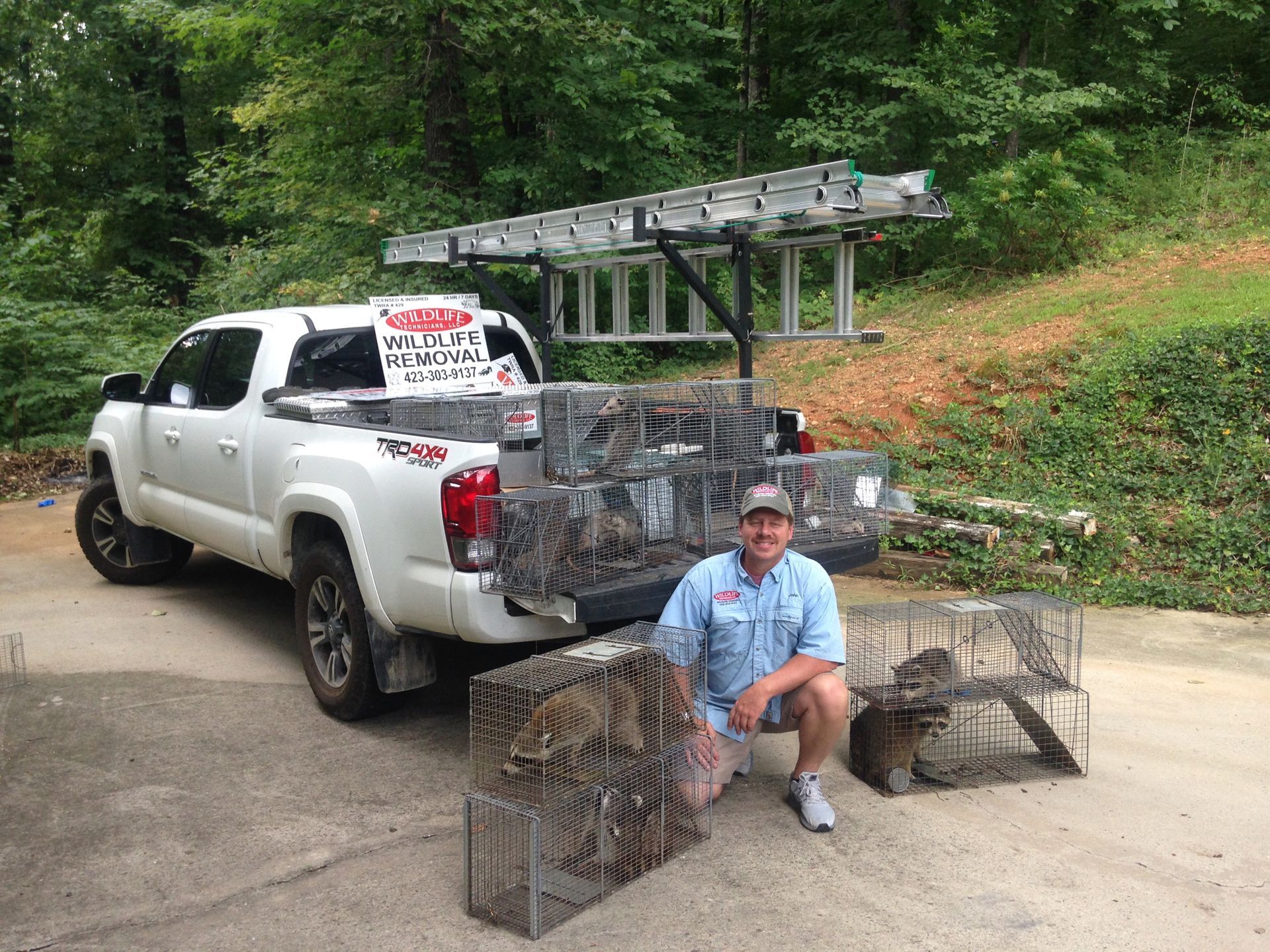 Man kneels beside pickup truck loaded with wire cages and a ladder in a wooded driveway.