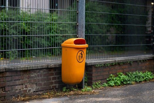 Orange public trash bin beside a brick wall and metal fence on a wet sidewalk.