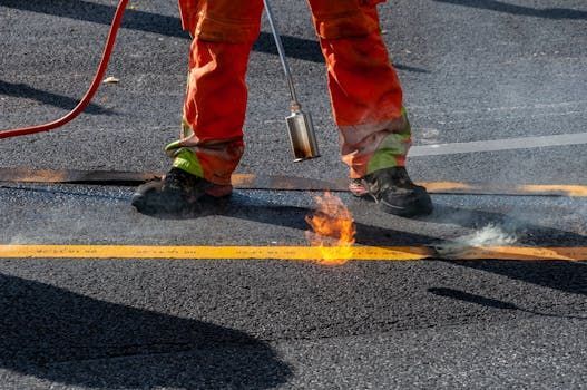 Worker in orange gear using a torch on asphalt, with flames and smoke on a road surface.