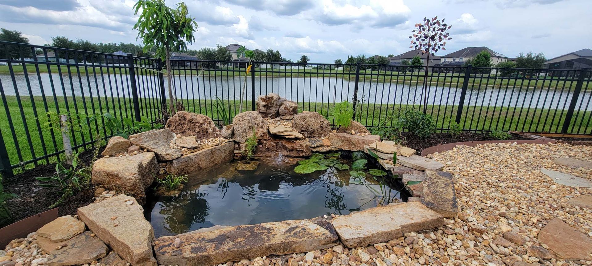 A small pond surrounded by rocks and a fence in a backyard.